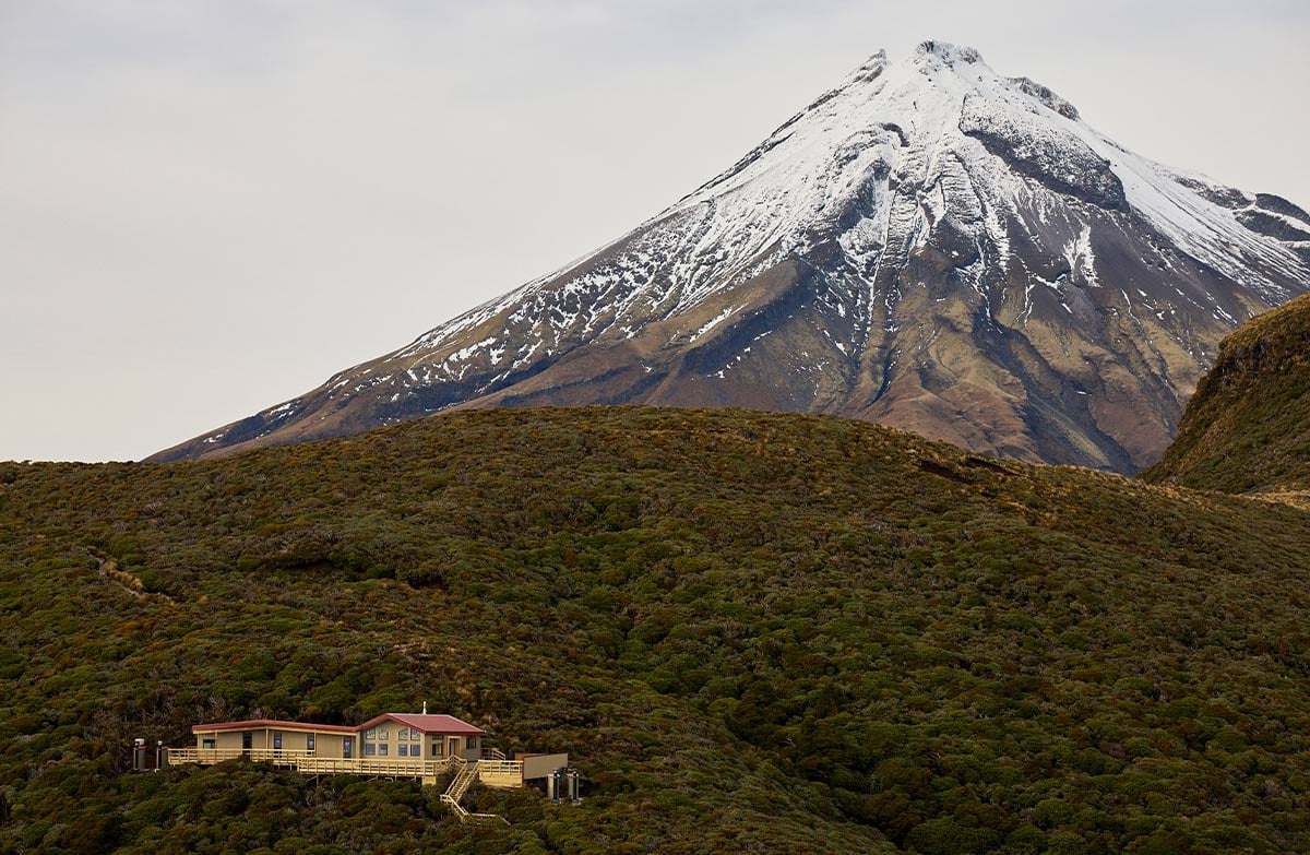 Snow capped Taranaki maunga looms over bush covered hills and a beige hut with a red roof sits nestled on a rise. 
