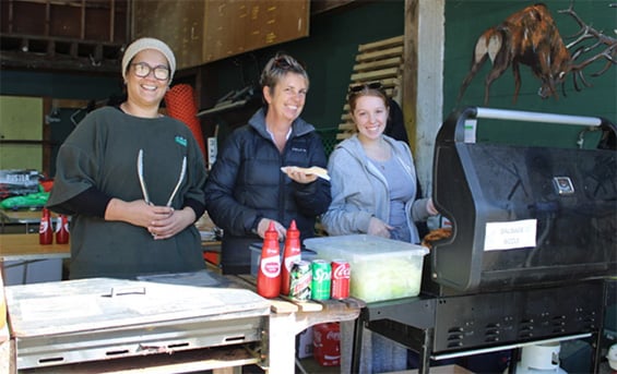 Three people stand behind a BBQ ready to serve food.