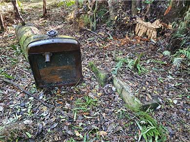 A rust covered metal cylinder connects to a metal box with radiator openings. It's in a forest