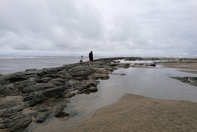 Two people walking across a rocky shore with a large rock pool in the foreground.