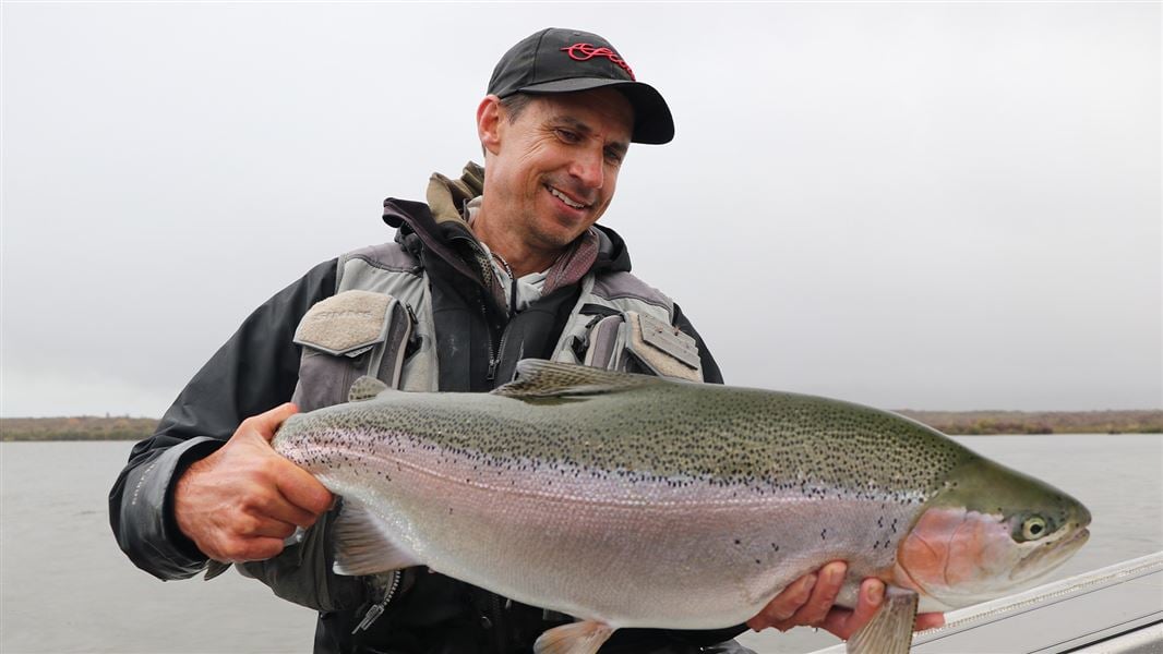 A man sitting on the side of a boat in the water holding a massive trout and grinning. 
