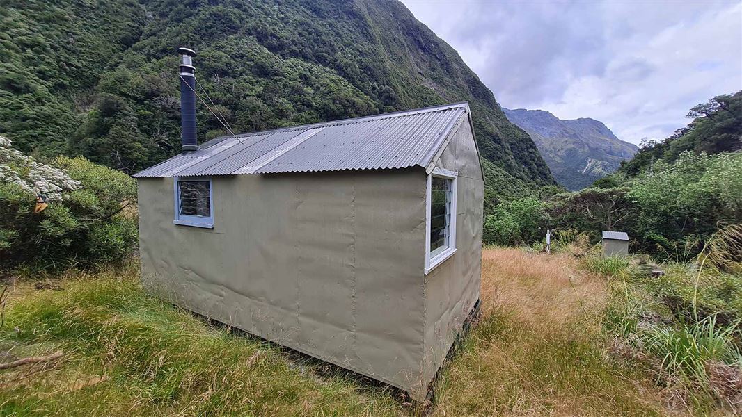 View from behind a small beige hut looking out over bush and distant mountains. 