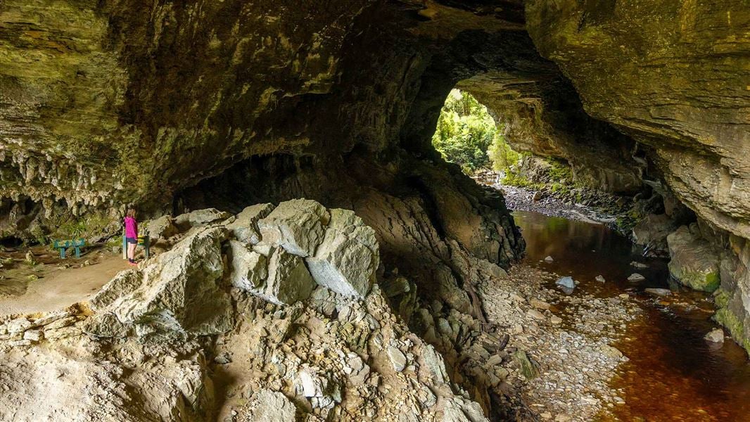 Large limestone archway with a stream flowing through it and an opening with native forest beyond. 