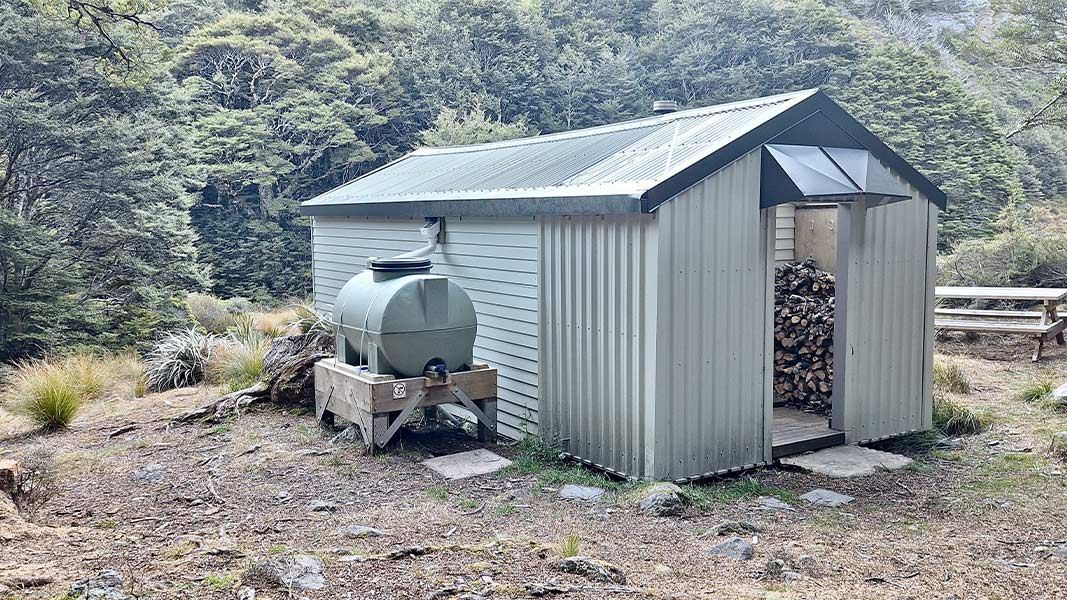 Ellis Hut a corrugated-metal hut in a bush clearing, with an open doorway showing stacked firewood inside and a green water tank mounted on a wooden stand beside it; picnic benches and dense forest in the background. 