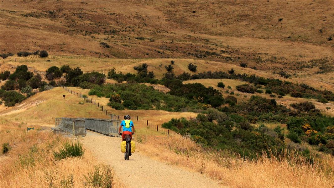 A lone cyclist approaches mountains along a gravel path.