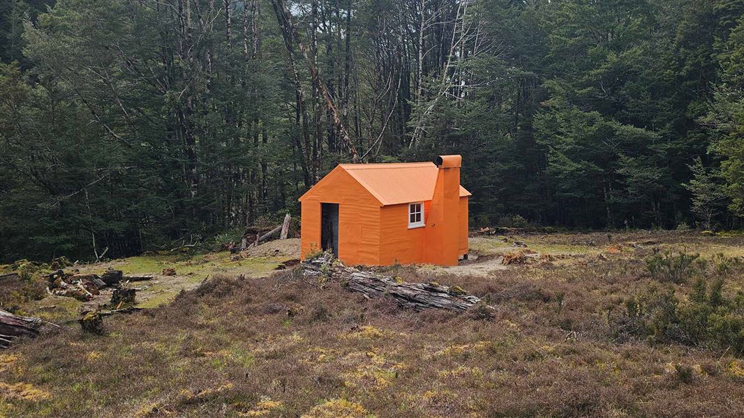 A small fully range hut sits on flat ground with tall trees behind it. 