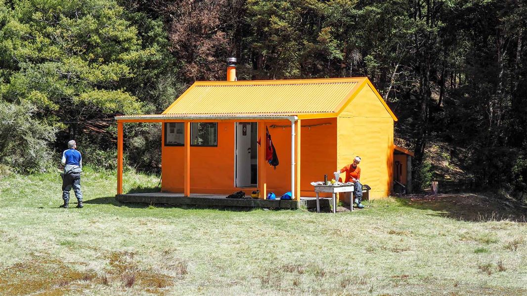 A small orange hut with huge trees behind it. Two people are outside, one sits at a table in the sun, the other is dressed in blue and facing the trees. 