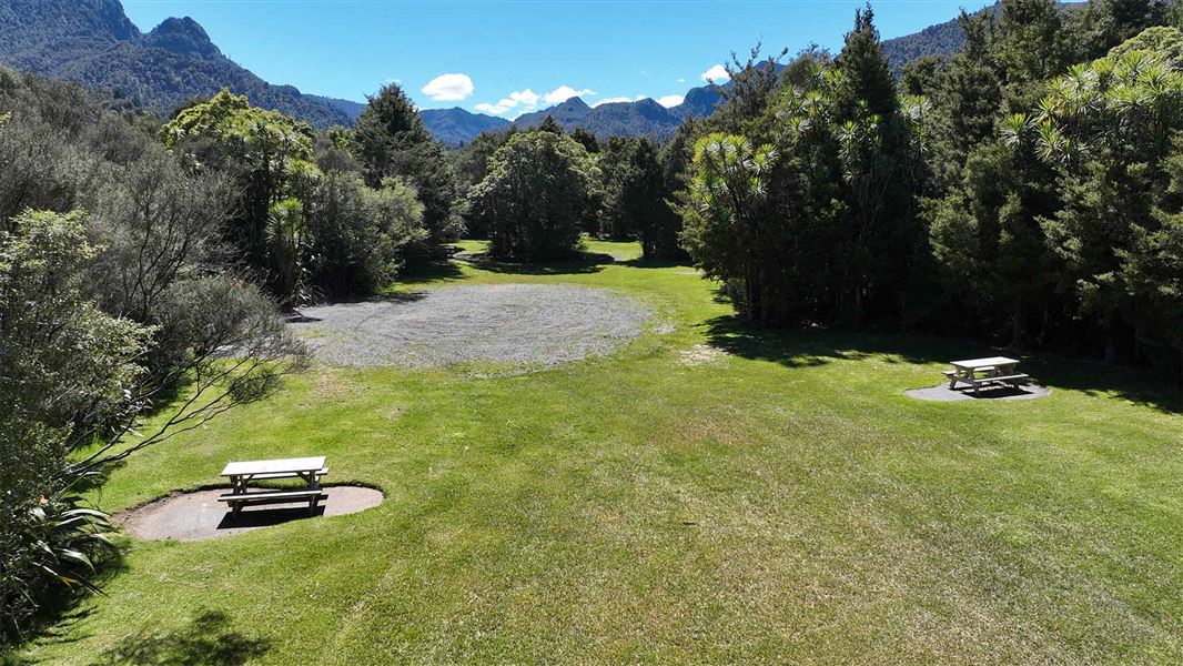 A flat grass area with picnic tables surrounded by bush and fauna covered mountains. 