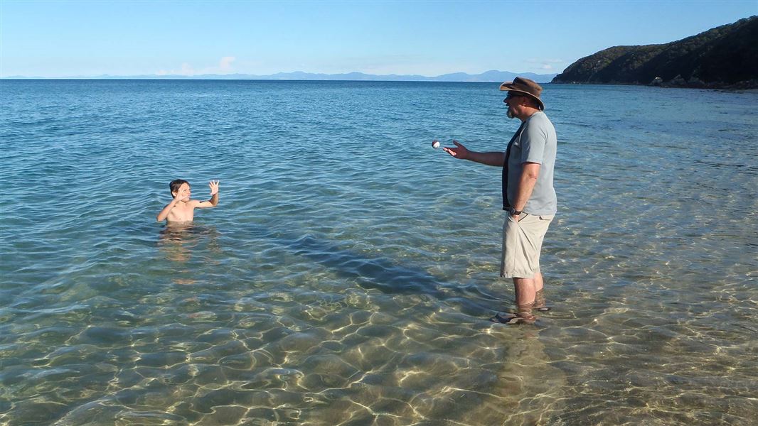 A man standing in clear, shallow coastal water holding a small ball, facing a child chest-deep in the water with raised hands. Calm sea extends to the horizon, with a forested headland visible on the right and a clear blue sky overhead.