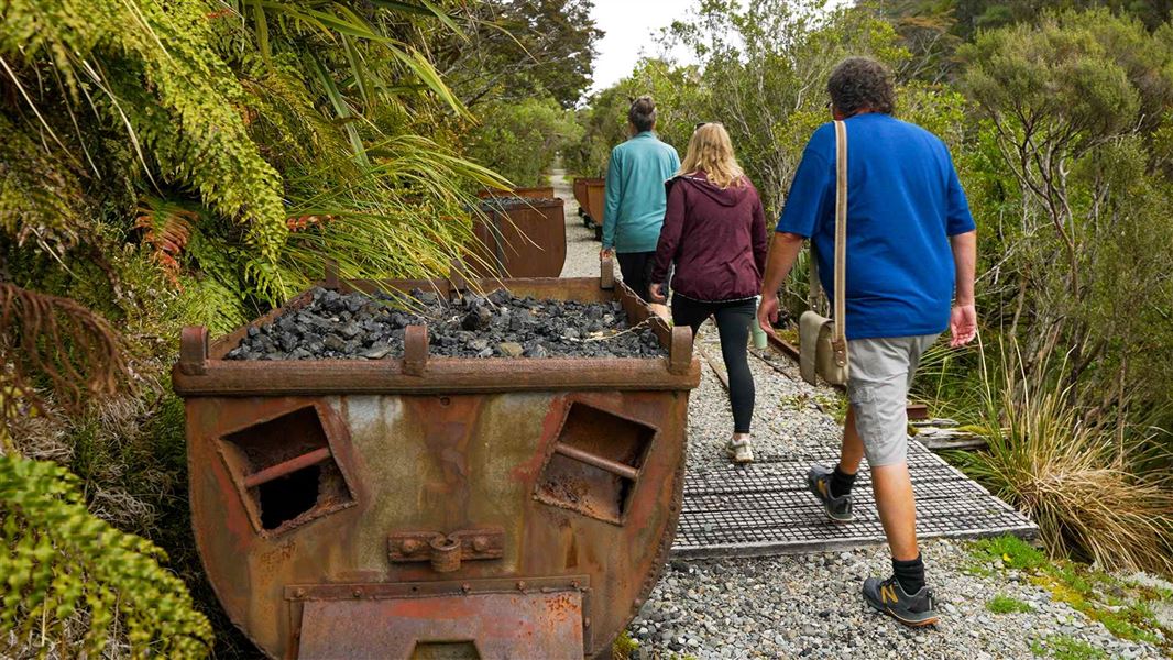 Three walkers pass small rusty wagons filled with coal on a gravel track through small subalpine shrubs and trees.