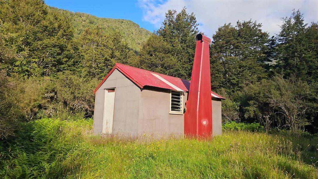 A small beige hut with a red chimney and roof, with bush behind. 