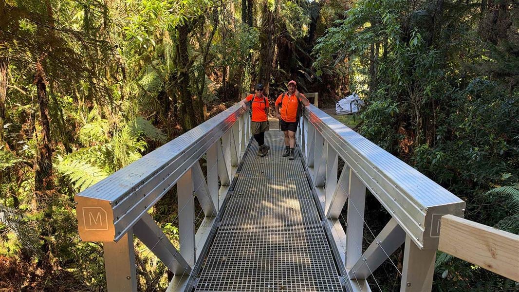 Two people in bright orange clothing pose on a wooden bridge et in the bush. 
