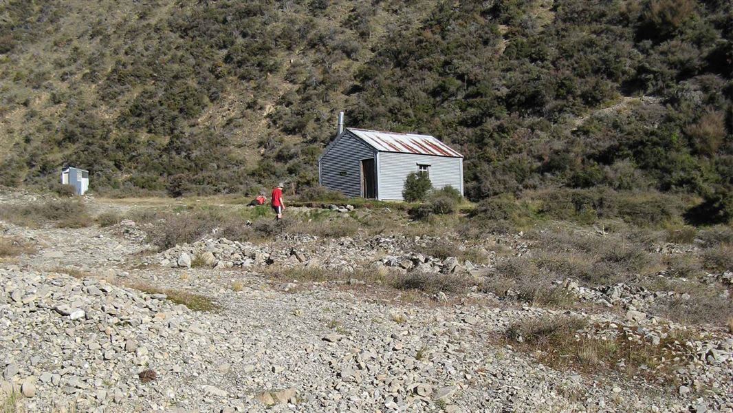 A tin-roofed hut sits on the rocky gravel near a river bank.