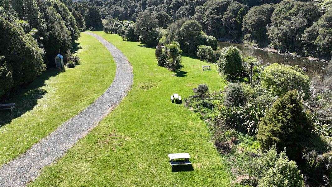 A gravel path runs through a grassy are with picnic tables, beside a bush lined stream. 