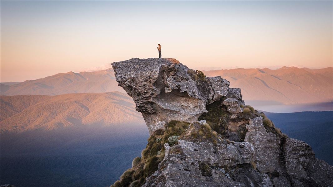 A huge rock shaped line a wave, with a person standing on top of it taking in the vast view of rolling reddy brown mountains. 