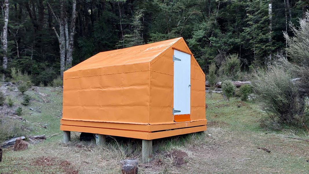 A small orange hut on piles, with a white door, sits on flat grass, with tall trees up behind it. 