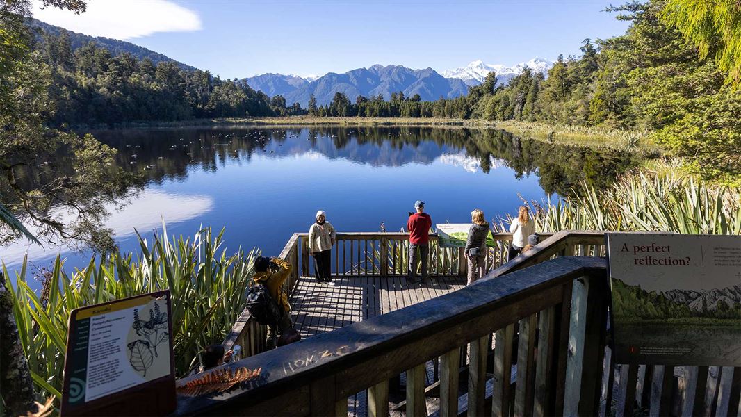 Visitors on a viewing platform overlooking the lake, with forest and mountain reflections in the still water.