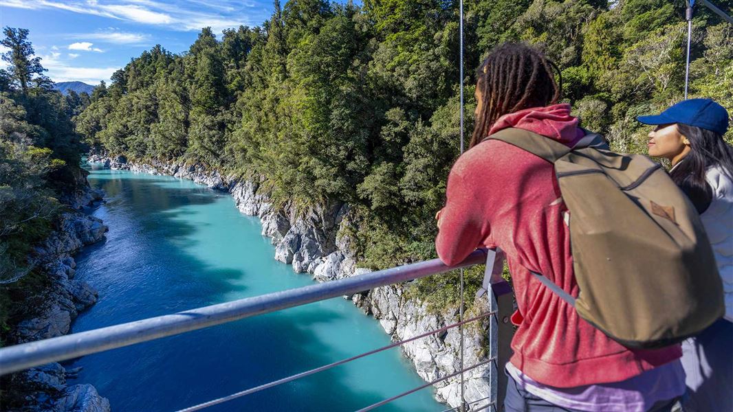 Two walkers on a bridge admire a wide river gorge with turquoise water and tall trees. 