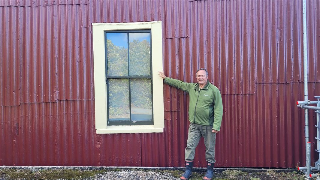 A man stands outside a corrugated iron clad wall leanng on a window frame.