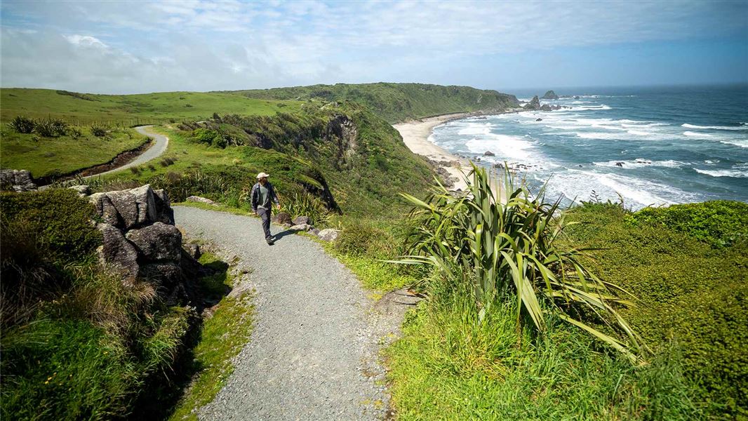 A walker on a gravel track high above a lush green coastline with wild surf beaches.