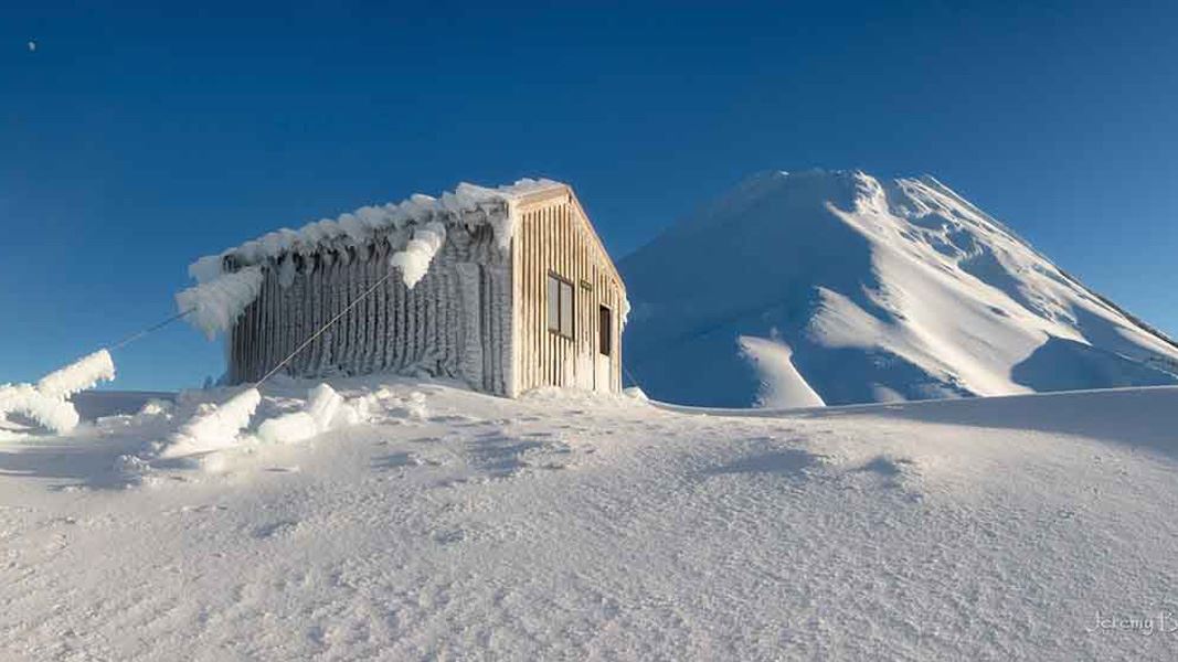 A small hut covered in windblasted snow with a large snow covered mountain behind it.
