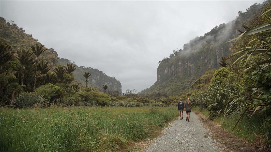 Two people walk down a gravel track with forest on either side and cliffs looming in the misty distance.