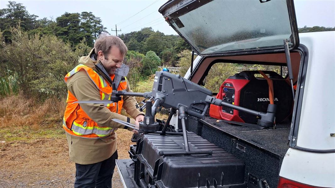 A man in hi vis vest stands in front of the open boot of his vehicle where a large drone sits on top of a container. 