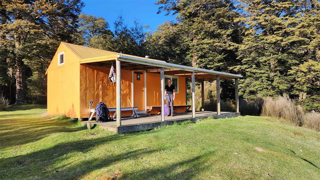 An orange hut with a covered wooden deck. A lady stands on the deck with a purple pack at her feet. 