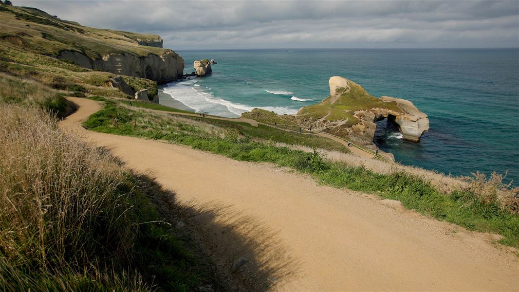 A gravel track winds down to a headland with a large natural rock archway spanning the sea.