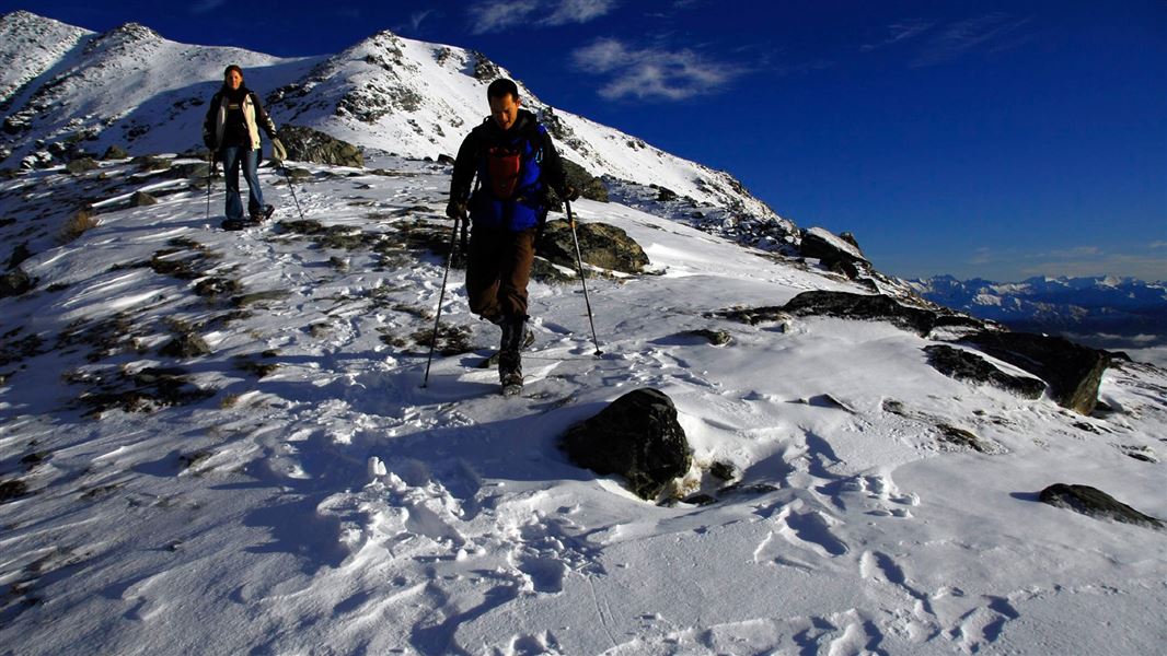 Two people carrying walking poles make their way down steep snowy mountain terrain. 