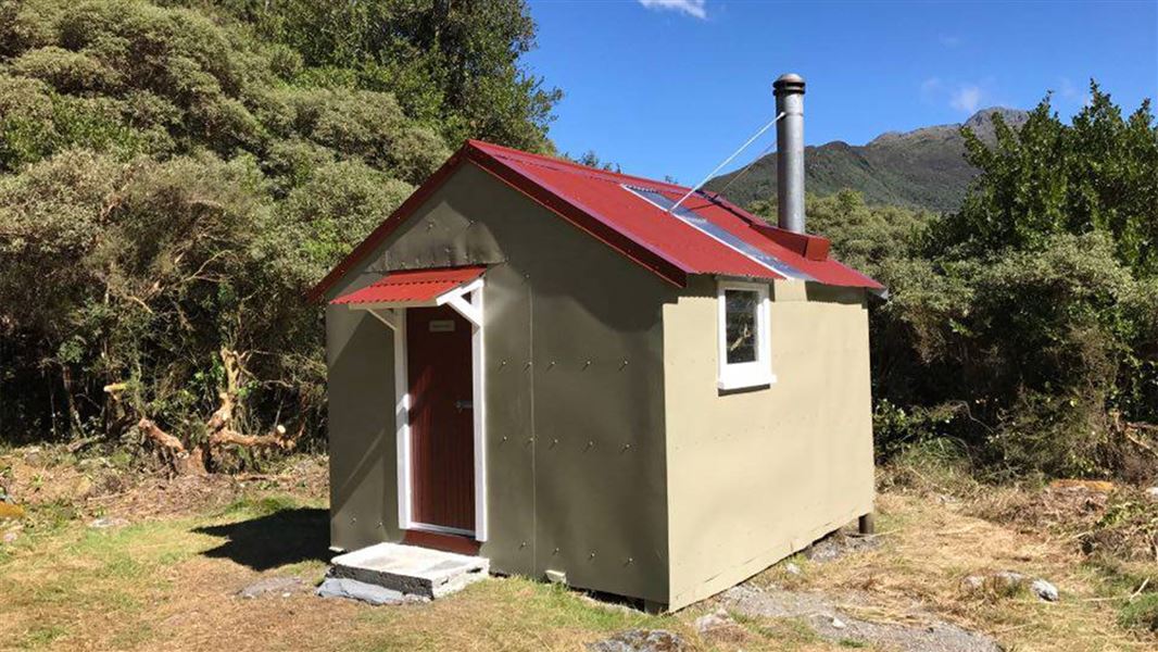 A small beige hut with a red roof and red door with a small concrete stoop, set in front of bush and hills. 