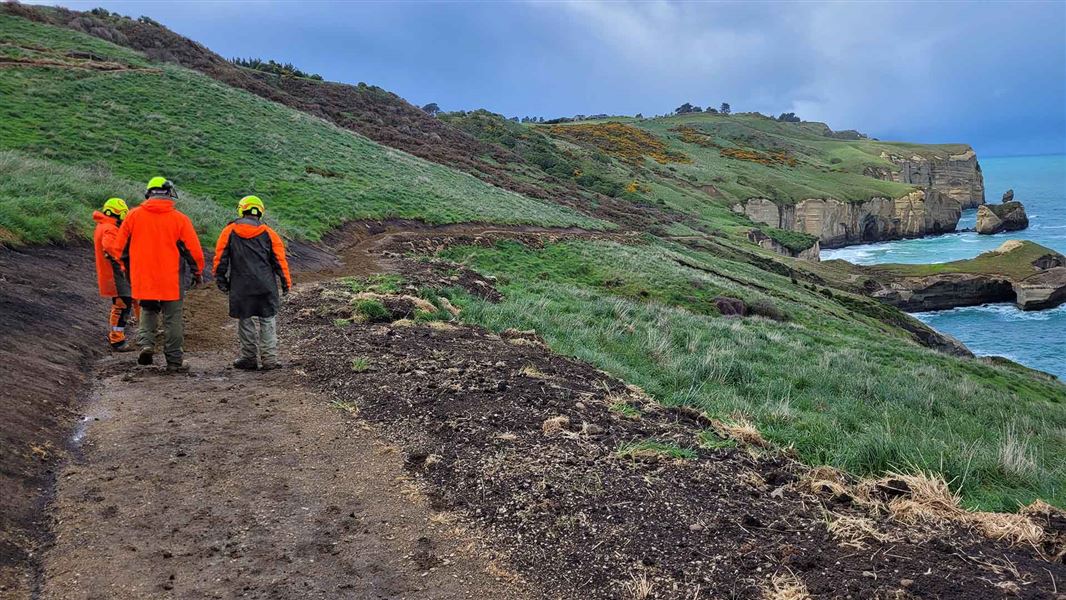 Three works in hi viz walk along a muddy path on cliffs above the sea.