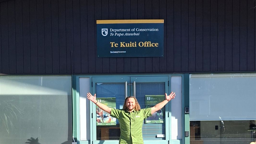 A DOC staff member in green uniform with his arms thrown out expansively. He stands in front of a DOC office with a Te Kuiti  Office sign attached to it. 