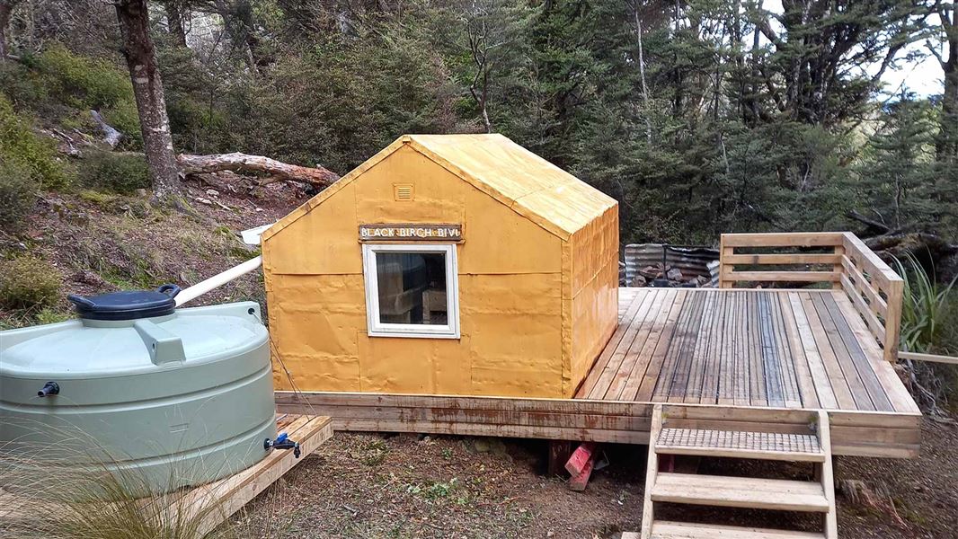 A small orange bivouac with a deck. A green plastic water tank sits to the left of it. 