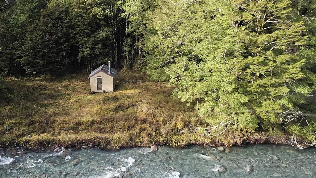 A tiny beige hut positioned on grass above a river with bush behind. 