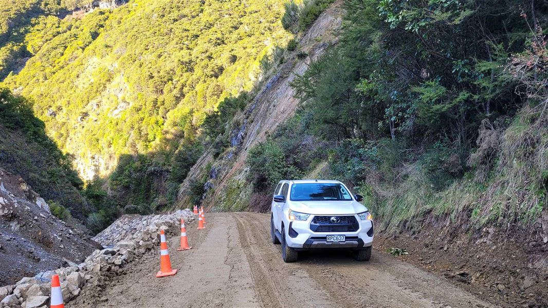 A white SUV on a dirt gravel road with road cones to the left and banked up rocks alongside a stream. 