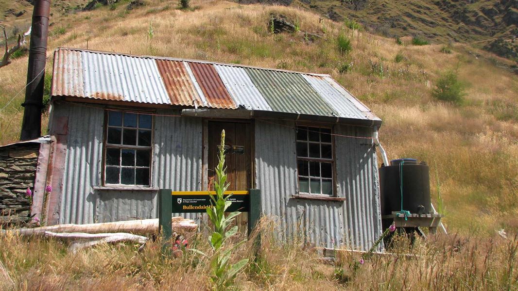 A corrugated tin hut with an external stone firebox and brown chimney, a small green water tank on the right of the building. Set at the bottom of a big wheat coloured hill. 