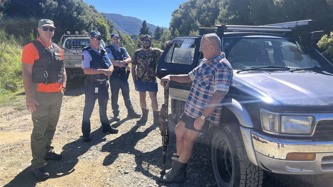 A group of people, including two uniformed police officers, stand beside a couple of 4WD vehicles in a wooded setting.