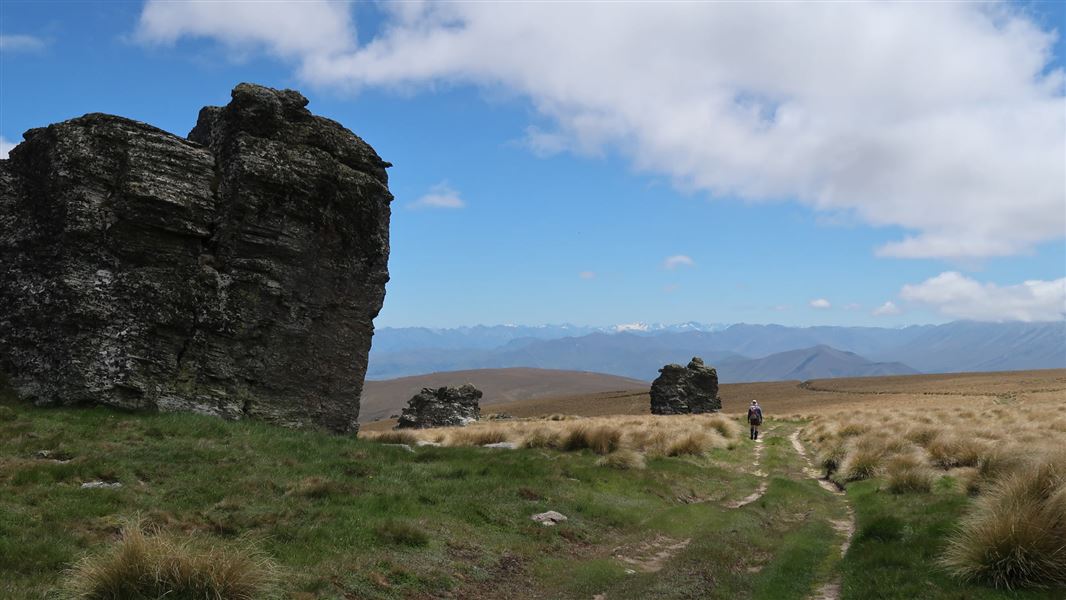 Large rock outcrops beside a grassy 4WD track crossing open tussock-covered terrain. A person is walking on the track near a smaller rock formation in the middle distance, with mountain ranges visible on the horizon beneath a partly cloudy sky.