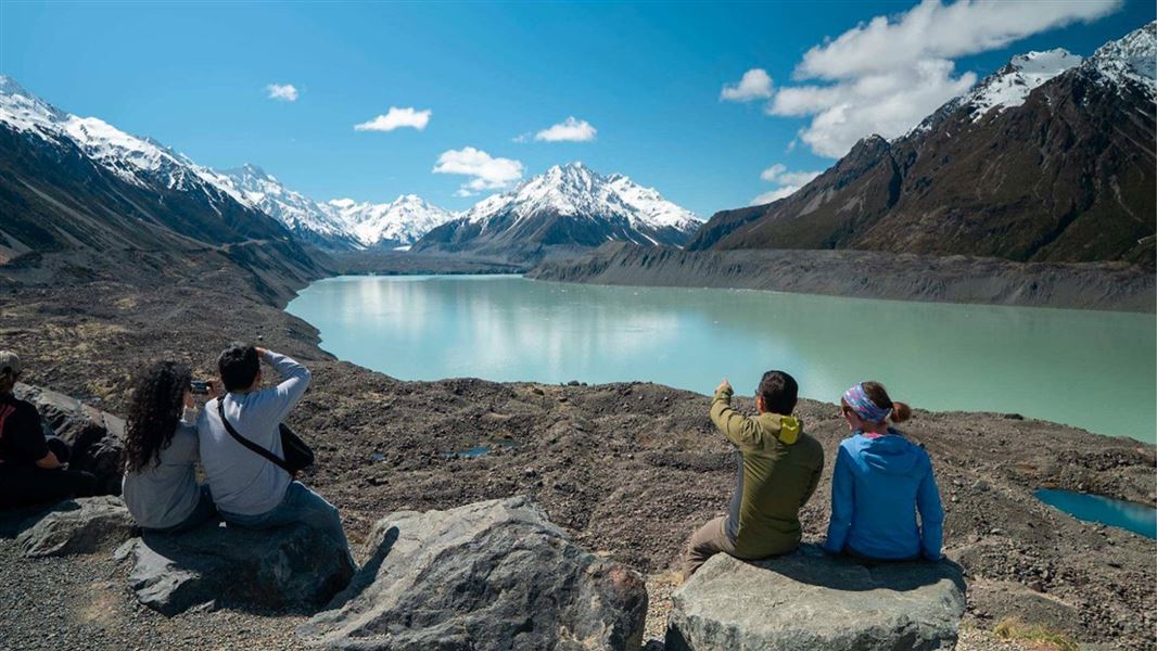 Foru people sit on rocks facing a flat glacial lake with Aoraki Mount Cook in the background covered in snow. 