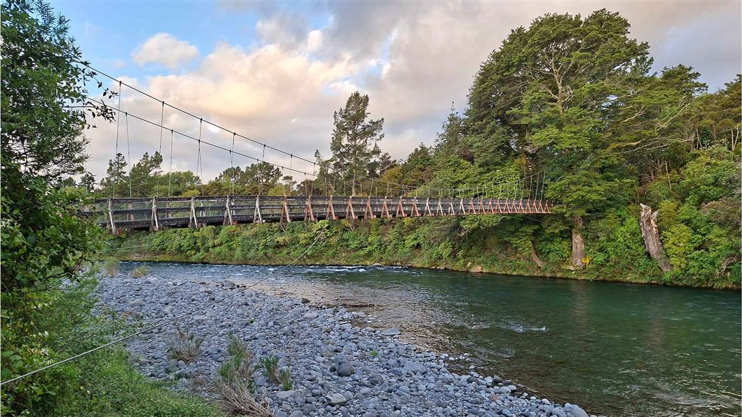 An aqua green river with a rocky beach sits beneath a swingbridge spanning forest. 