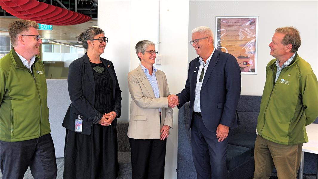 Two people in business attire shake hands while three others watch on. Everyone smiles.