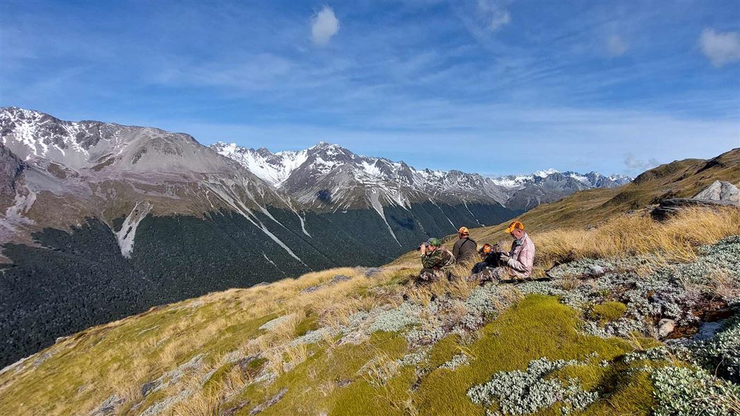 Group of hunters gathered outdoors in a rural New Zealand landscape during a managed hunt, with rolling hills and vegetation visible in the background.