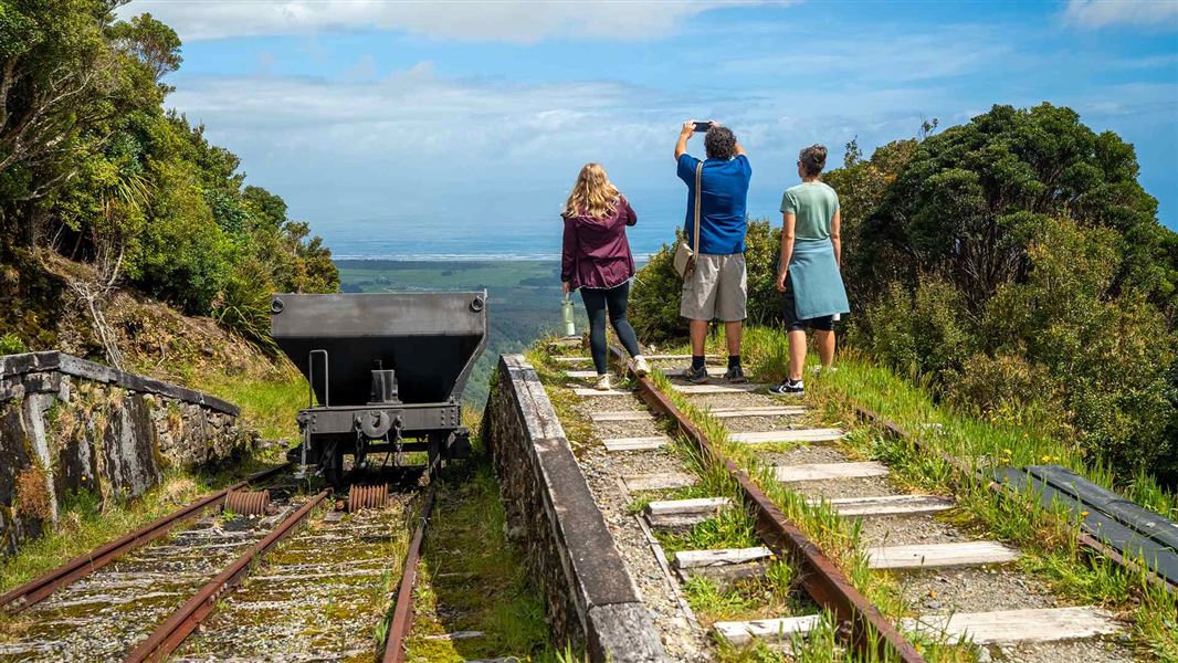Three visitors beside an old coal wagon on a tramline plunging steeply downhill photograph sweeping coastal views.