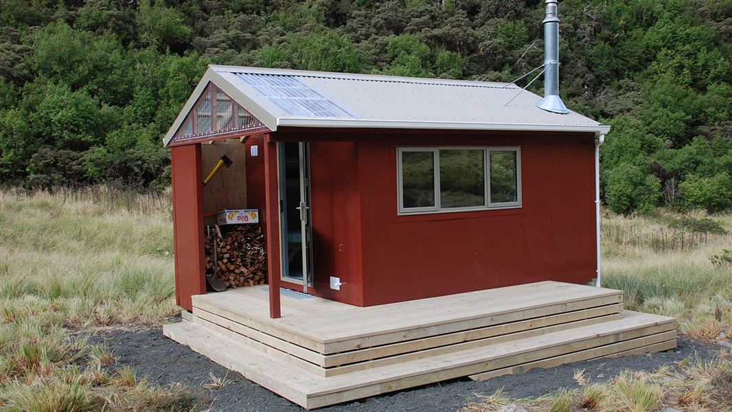 A small reddish brown hut with a small pale wood deck sitting on a gravel pad amongst grass and bush. 