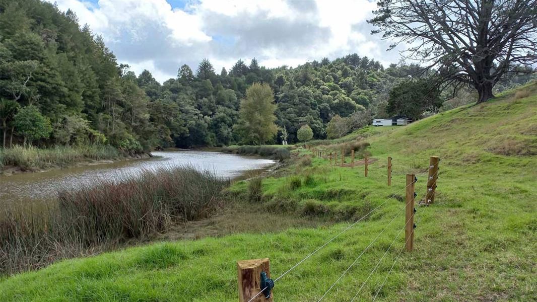 Grassy landscape with a brown river running through it and a fence visible beside it. 