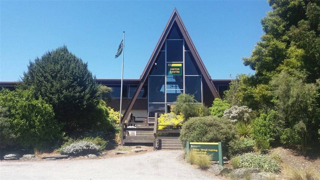 The A-framed DOC visitor centre located in Wānaka. It has mostly windows on the triangular apex and is surrounded by foliage. 