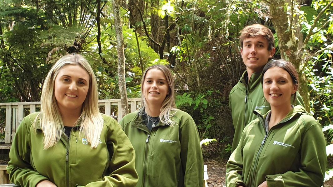 Four young people standing in DOC uniforms in the bush on a sunny day. 