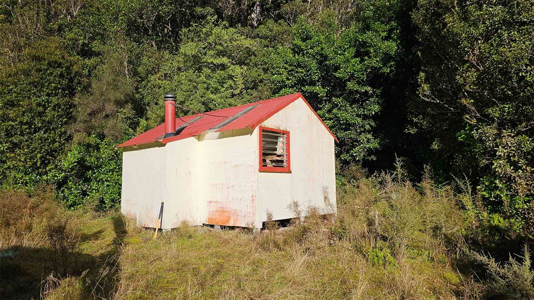 A corrugated beige hut with a red roof and chinmey sitting on grass with large trees behind it. 