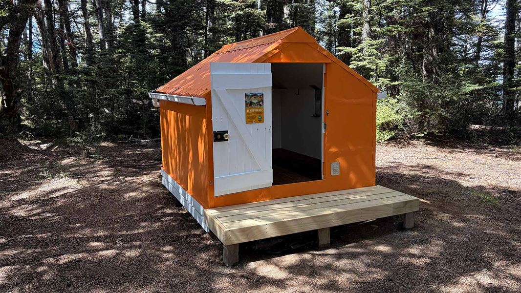 A small orange bivvy with the door open, the door interior is white. The bivvy sits on foundations and has a small deck. It is in a forest clearing with tall trees behind it. 